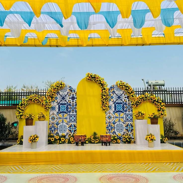 A brightly colored outdoor event stage setup for a traditional Indian ceremony, featuring a backdrop of vertical fabric drapes in yellow, magenta, and purple. The top of the backdrop is lined with five decorative, multi-colored Rajasthani embroidered umbrellas. Diamond-shaped paper kites and hanging tassels decorate the fabric. In the foreground, a central low bench wrapped in yellow fabric sits between two gold pots on stands, from which thick garlands of yellow marigold flowers spill out onto the grass.