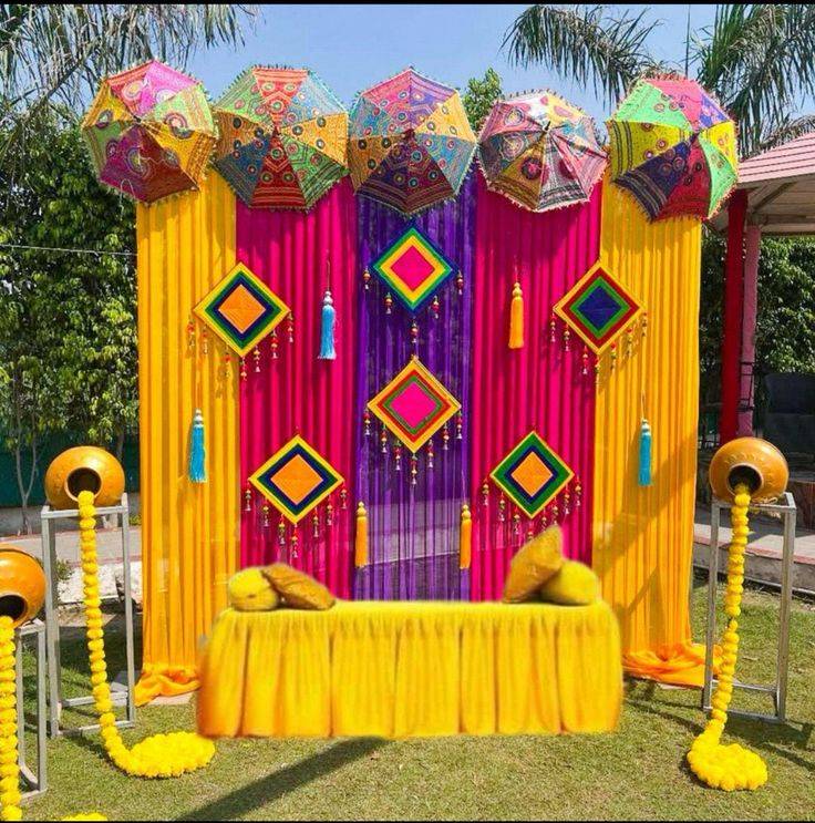 A brightly colored outdoor event stage setup for a traditional Indian ceremony, featuring a backdrop of vertical fabric drapes in yellow, magenta, and purple. The top of the backdrop is lined with five decorative, multi-colored Rajasthani embroidered umbrellas. Diamond-shaped paper kites and hanging tassels decorate the fabric. In the foreground, a central low bench wrapped in yellow fabric sits between two gold pots on stands, from which thick garlands of yellow marigold flowers spill out onto the grass.